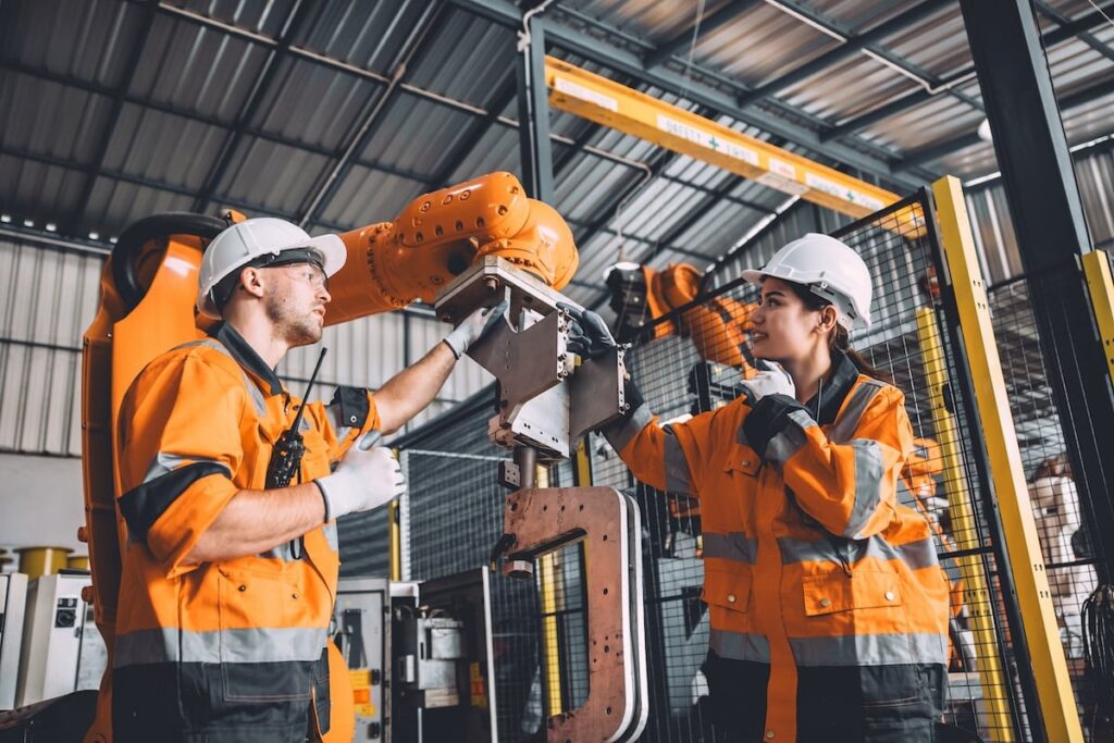 Deux techniciens en tenue de sécurité orange, portant des casques et des gants, travaillent sur un bras robotique dans un environnement industriel. Ils inspectent et ajustent l'équipement, illustrant une scène de maintenance et de prévention des risques en milieu de travail.